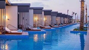 Uma área de piscina moderna de hotel com várias espreguiçadeiras e cadeiras de banho ao longo da borda, cercada por palmeiras e um céu límpido.