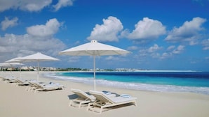 Uma praia com guarda-sóis brancos e espreguiçadeiras, céu azul límpido e mar calmo.