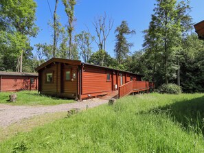 Une cabane en bois dotée d’un porche et d’une petite fenêtre, entourée de grands arbres et d’une zone herbeuse.