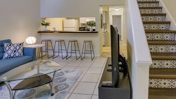 A living room with a glass coffee table, a sofa, and a TV. There are three stools and a small table with a lamp. A kitchen is visible in the background with a refrigerator and a plant. - 4 Mi to Dtwn: Pet-Friendly Abode in Tallahassee! (Tallahassee)