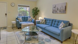 A living room with a blue sofa, a glass coffee table, a patterned rug, a cityscape painting, and a potted plant. - 4 Mi to Dtwn: Pet-Friendly Abode in Tallahassee! (Tallahassee)