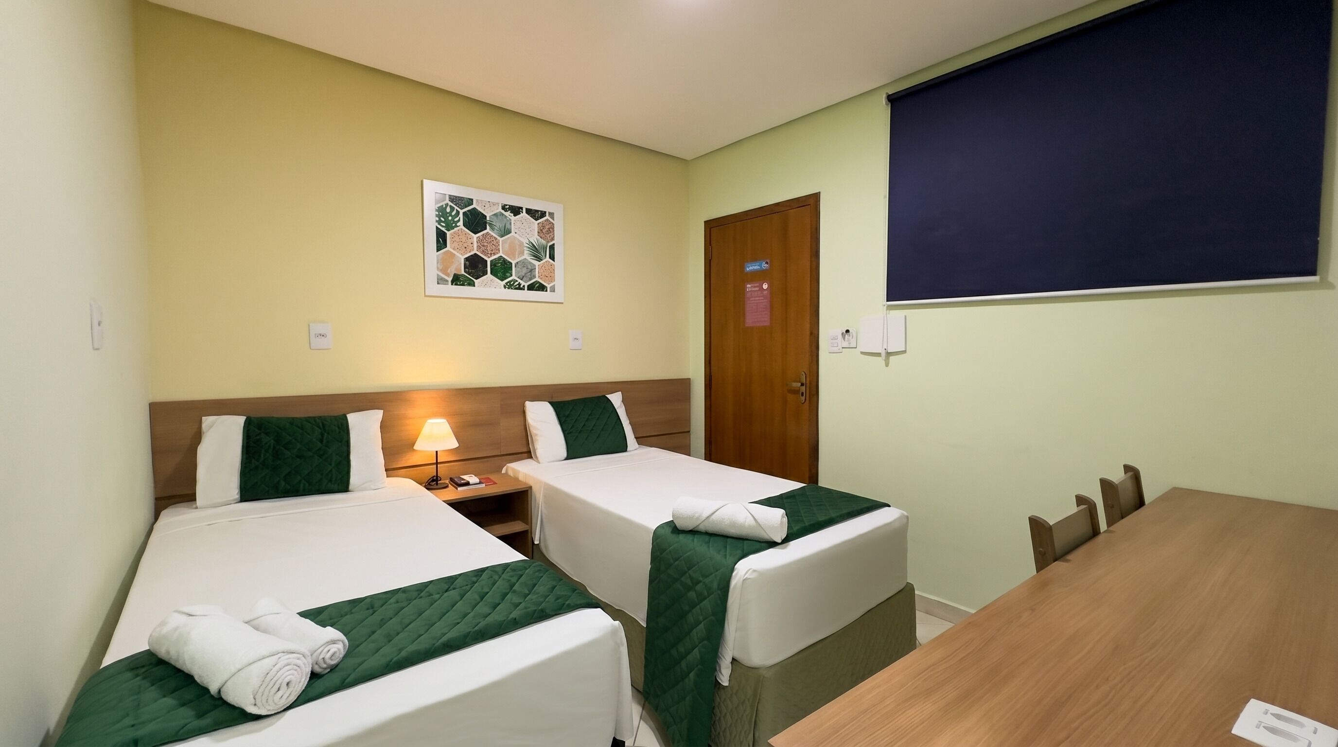 A hotel room with two beds, a wooden table, a wall-mounted picture, and a door.