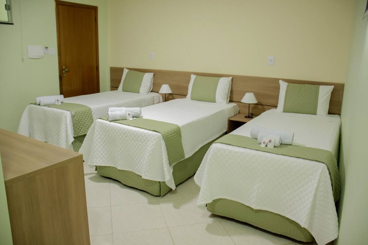 A hotel room with three single beds, each with a green bedspread and white pillows, a wooden nightstand with a lamp, and a wooden desk.