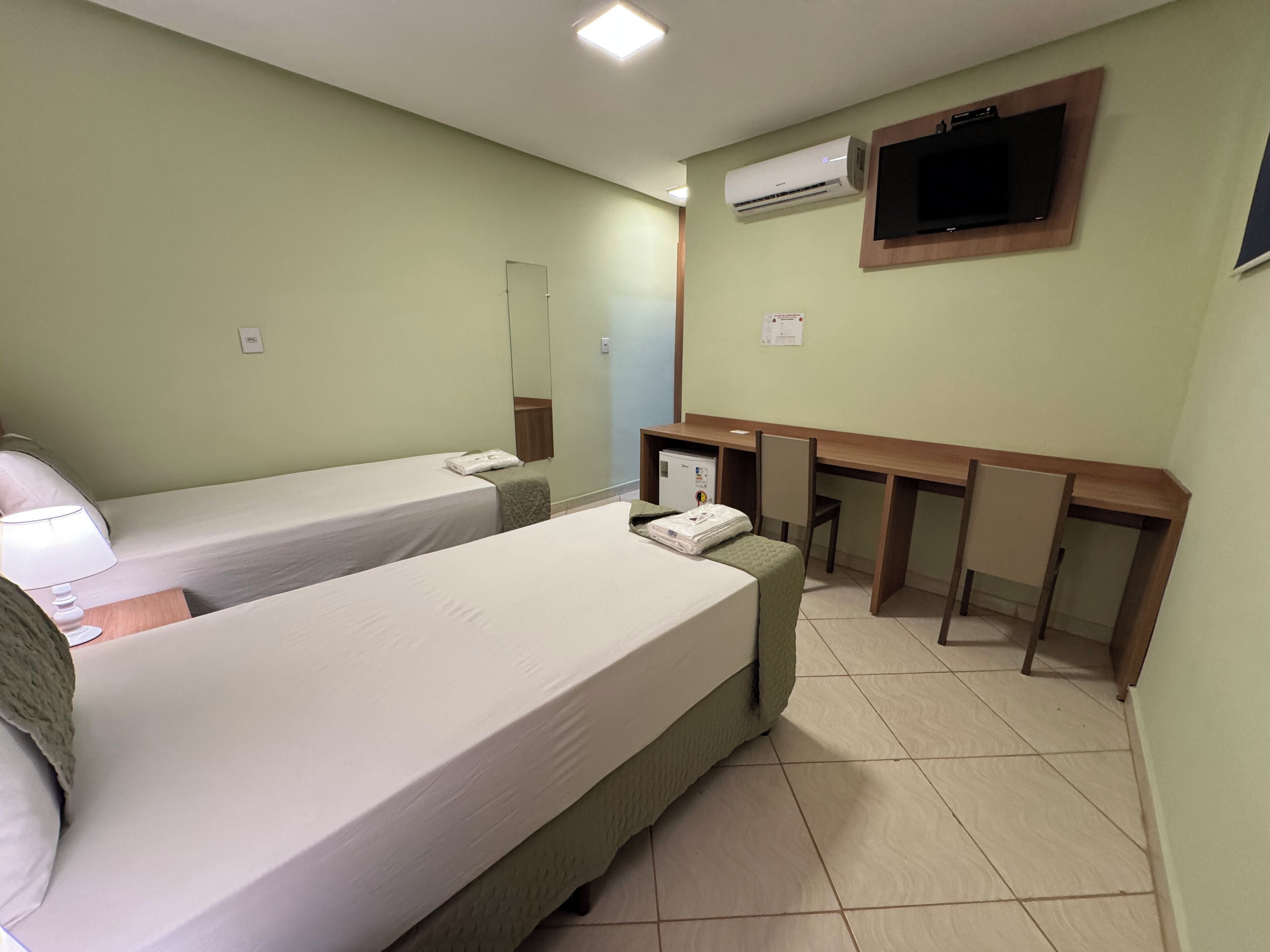 Une chambre d’hôtel avec deux lits simples, un bureau avec une chaise, un téléviseur fixé au mur et un appareil de climatisation.