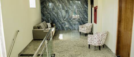 A lobby with a marble wall, a sofa, two patterned chairs, a wooden door, and a staircase with metal railings.