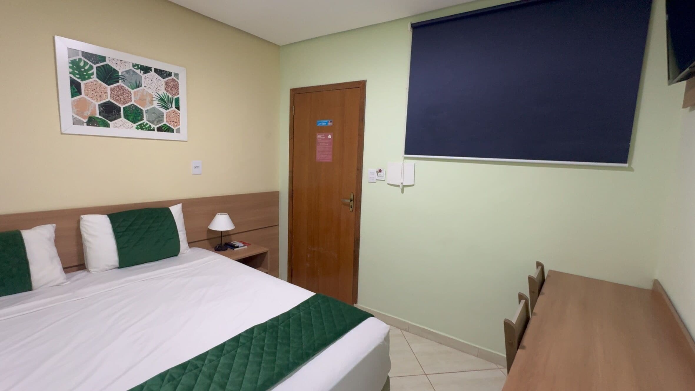 A hotel room with a bed, a desk, a chair, a wall-mounted TV, and a door with a sign.