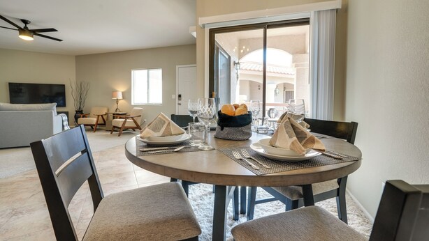 A dining area with a round table set for four, including plates, cutlery, and napkins. There are four chairs around the table, and a ceiling fan is visible above. The room has a large window with curtains, and a sofa is visible in the background.
