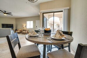 A dining area with a round table set for four, including plates, cutlery, and napkins. There are four chairs around the table, and a ceiling fan is visible above. The room has a large window with curtains, and a sofa is visible in the background. - Pool Access! 55 + Snowbird Retreat in Peoria (Peoria)