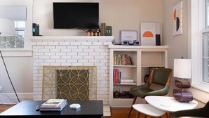 Un salón con chimenea de ladrillo blanco, un televisor de pantalla plana montado encima, una estantería con libros y objetos decorativos, un sillón verde, una mesita redonda y una lámpara.