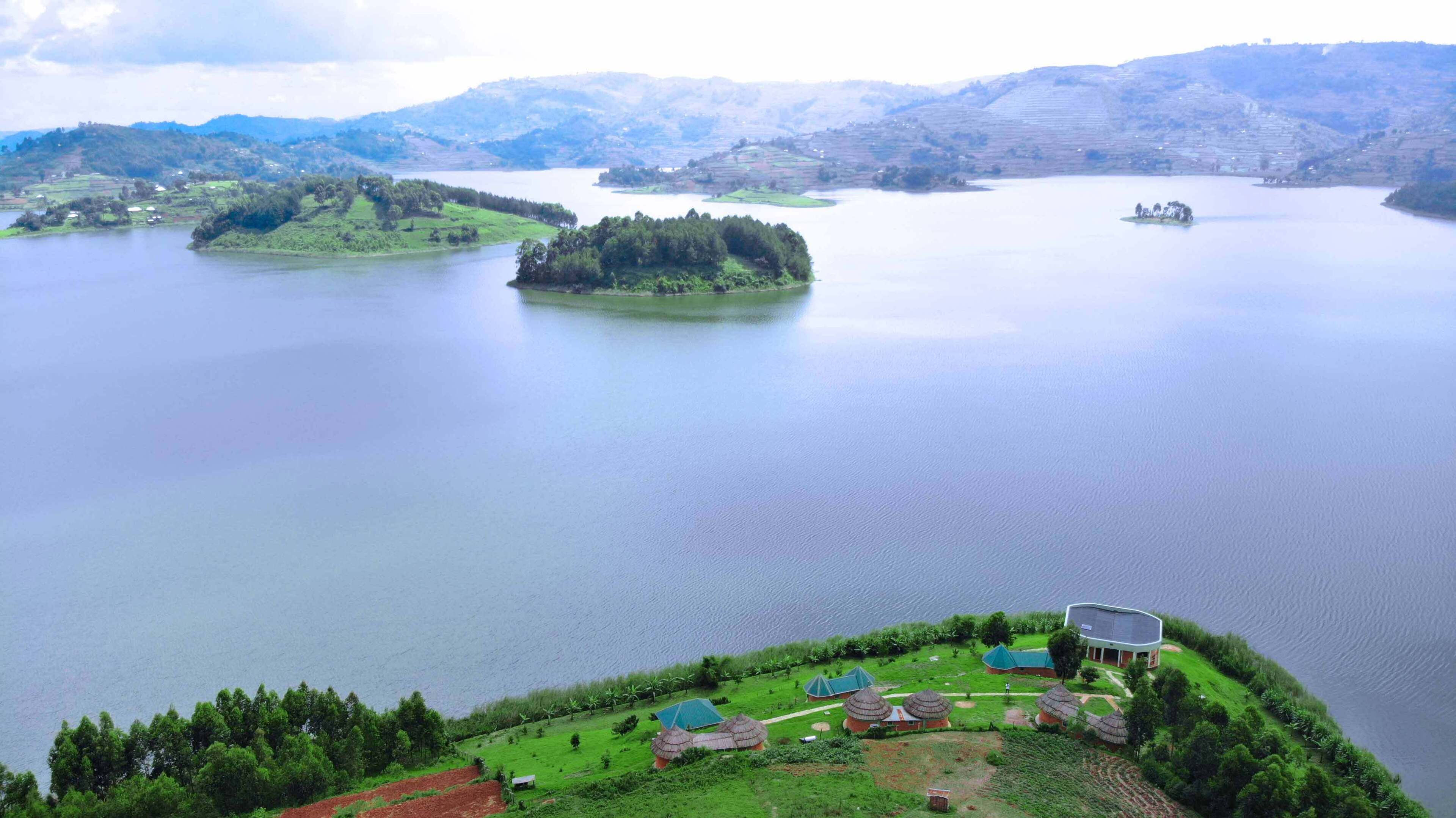A lakeside resort with multiple thatched-roof huts, a large building with a curved roof, and a view of the surrounding hills and water.