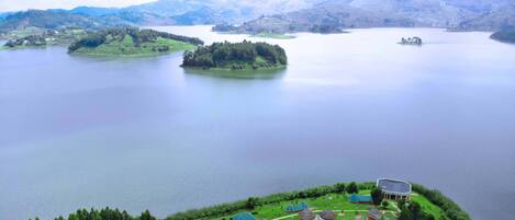 A lakeside resort with multiple thatched-roof huts, a large building with a curved roof, and a view of the surrounding hills and water.