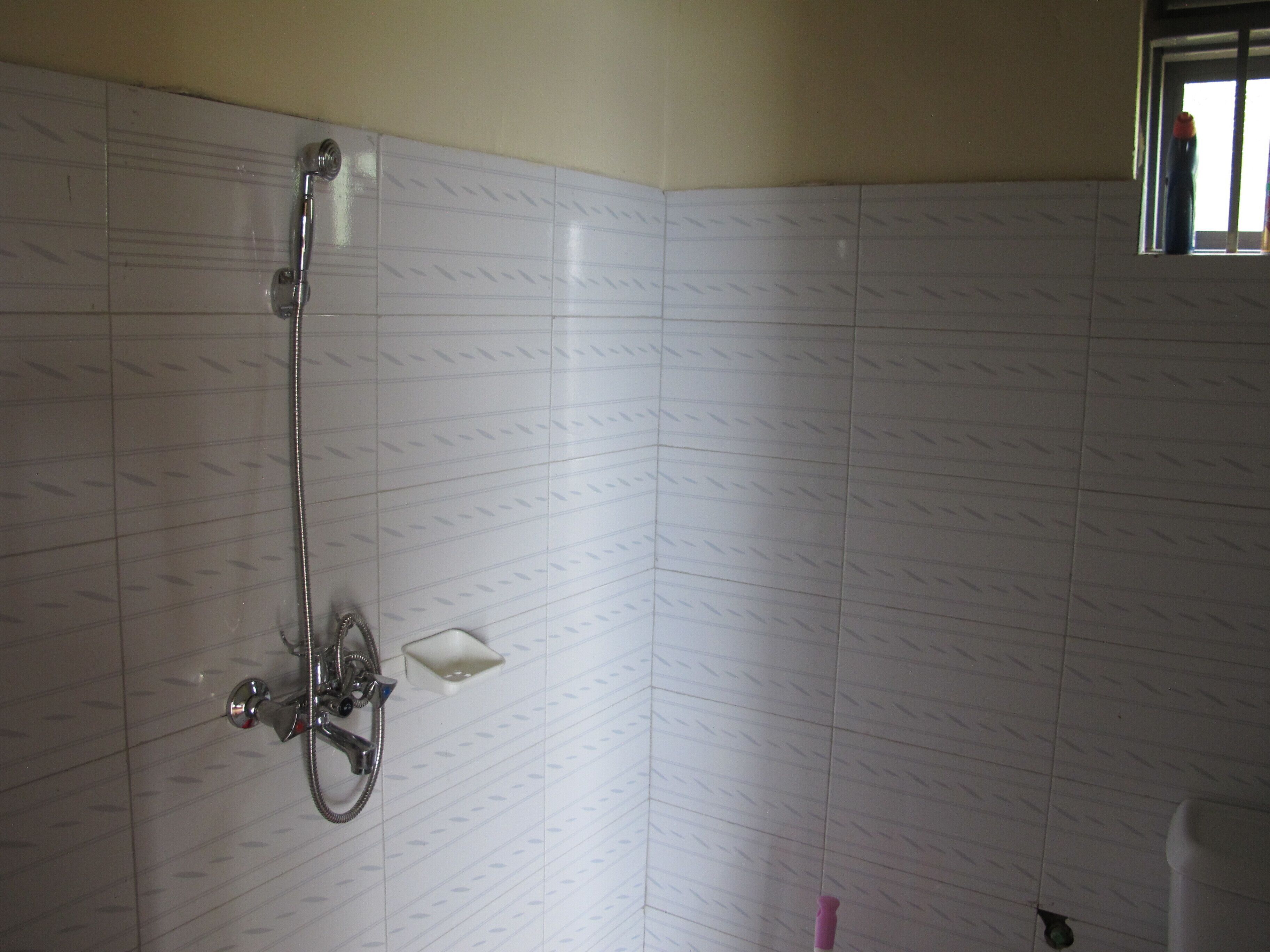 A bathroom with white tiled walls, a shower with a handheld showerhead, and a small white shelf.
