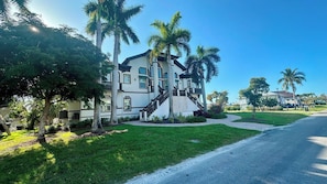 A large, two-story house with a prominent staircase leading to the entrance, surrounded by lush greenery and palm trees.