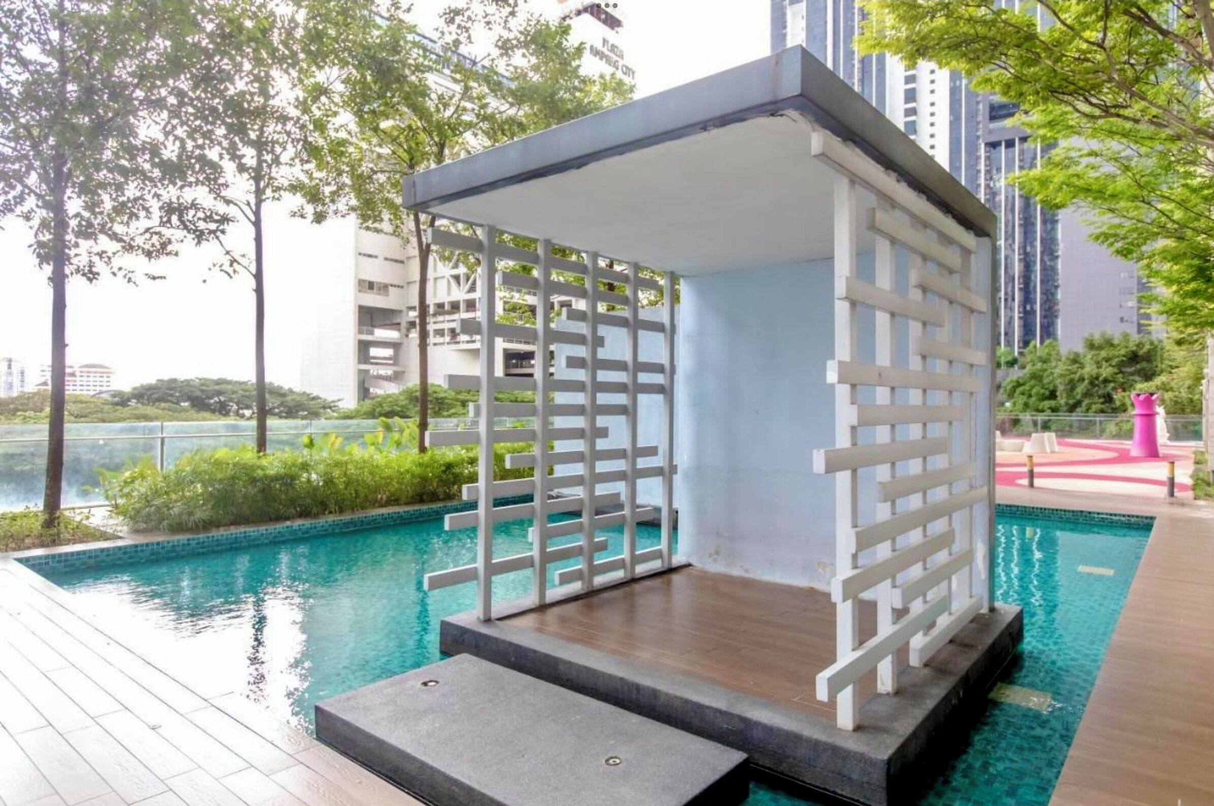 Un área de piscina moderna al aire libre con una zona de estar cubierta, rodeada de vegetación y edificios altos.