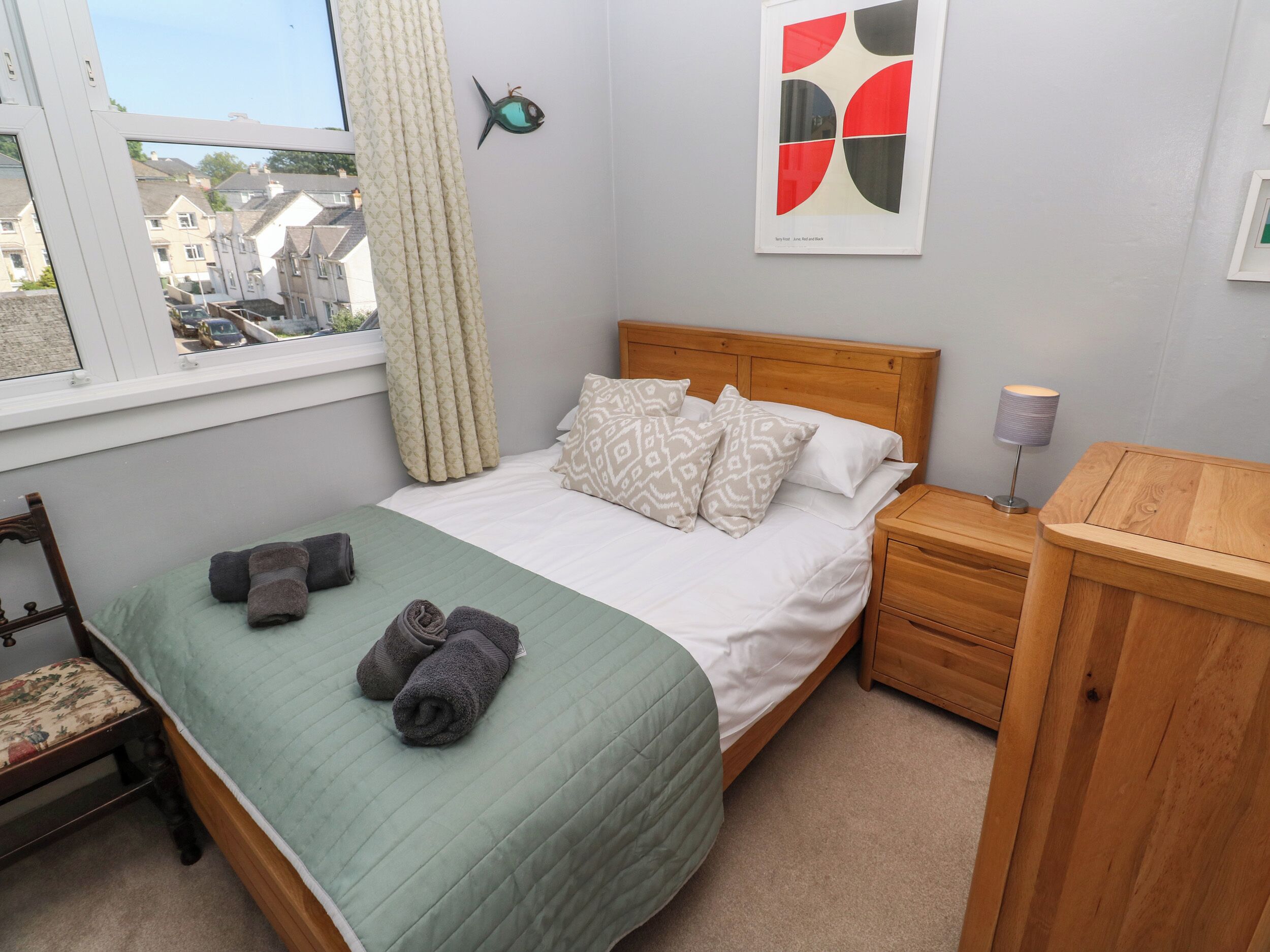 A neatly made bed with a green quilt, two rolled towels, and patterned pillows. A wooden nightstand with a lamp is beside the bed. A window with curtains is visible, and a fish-shaped wall decoration is above the bed.