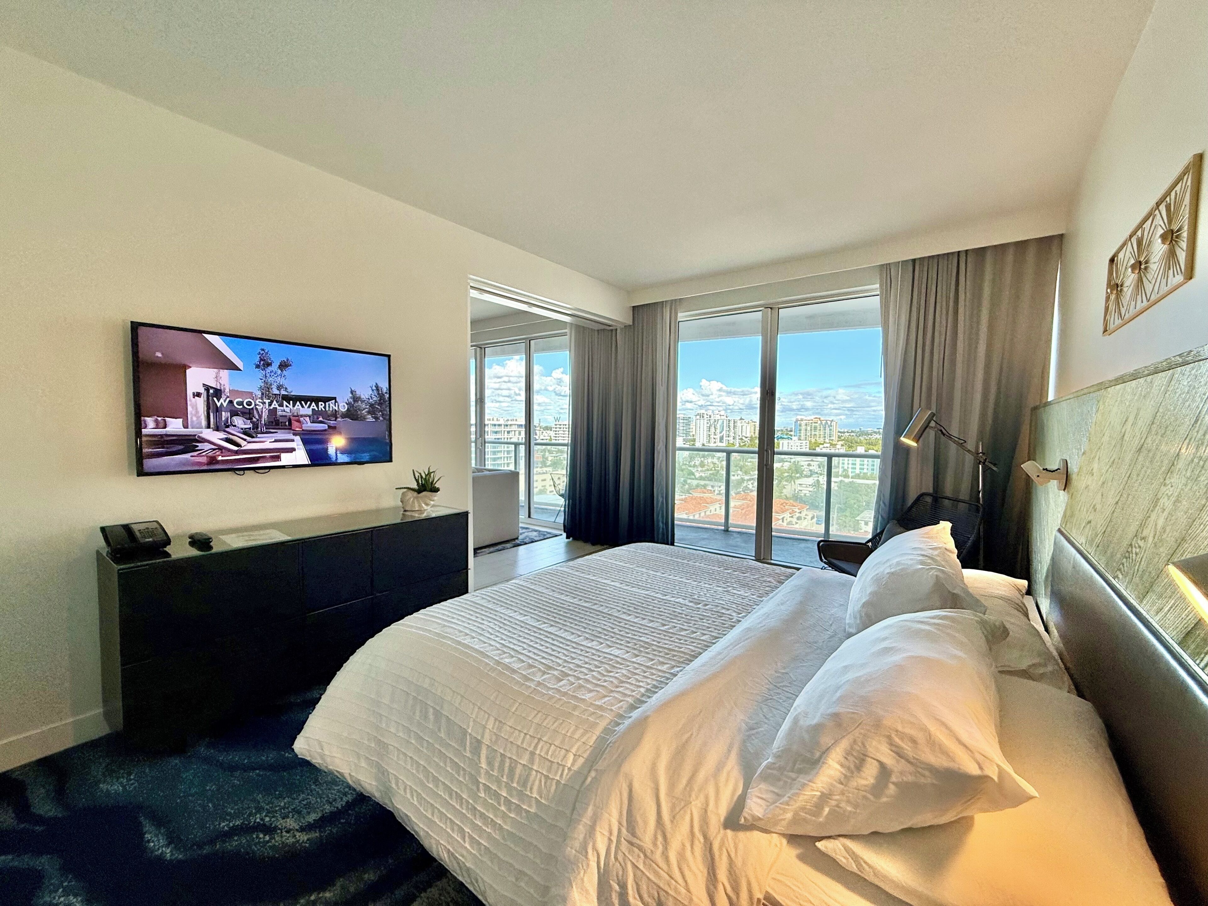 Hotellihuone, jossa on sänky, seinälle asennettu televisio, työpöytä puhelimineen ja suuri ikkuna, josta avautuu näkymä kaupunkiin.