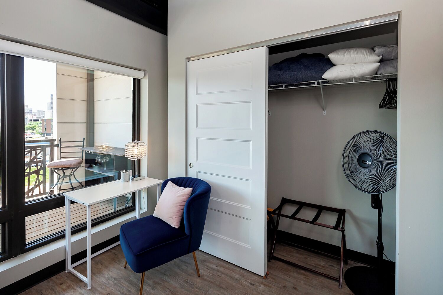 Une pièce comprenant un bureau, une chaise, un placard ouvert et un ventilateur sur pied.