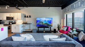 A modern living room with a grey sofa, a red chair, a flat-screen TV, and a kitchen area with white cabinets and a microwave. - The Cartier Lux DT Apt w/Skyline View (Des Moines)