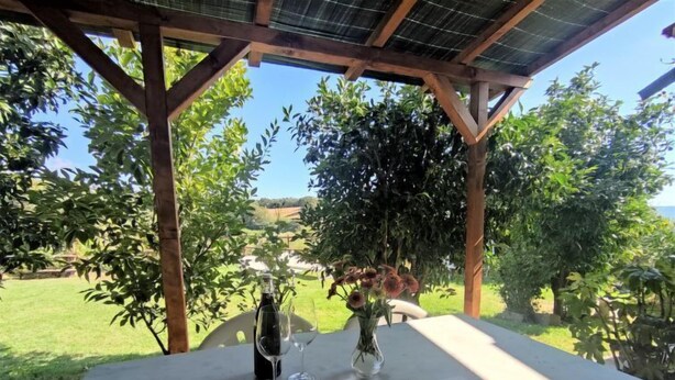 Une table installée à l’extérieur, avec un livre, une bouteille de vin et des verres, placée sous une pergola d’où l’on peut admirer un paysage verdoyant.
