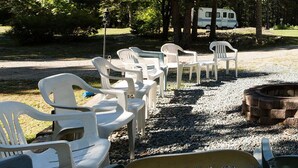 A campground with white plastic chairs arranged in rows, a fire pit, and a white RV parked among trees. - 3BedroomApt Pool CampFire PlayArea (Bar Harbor)