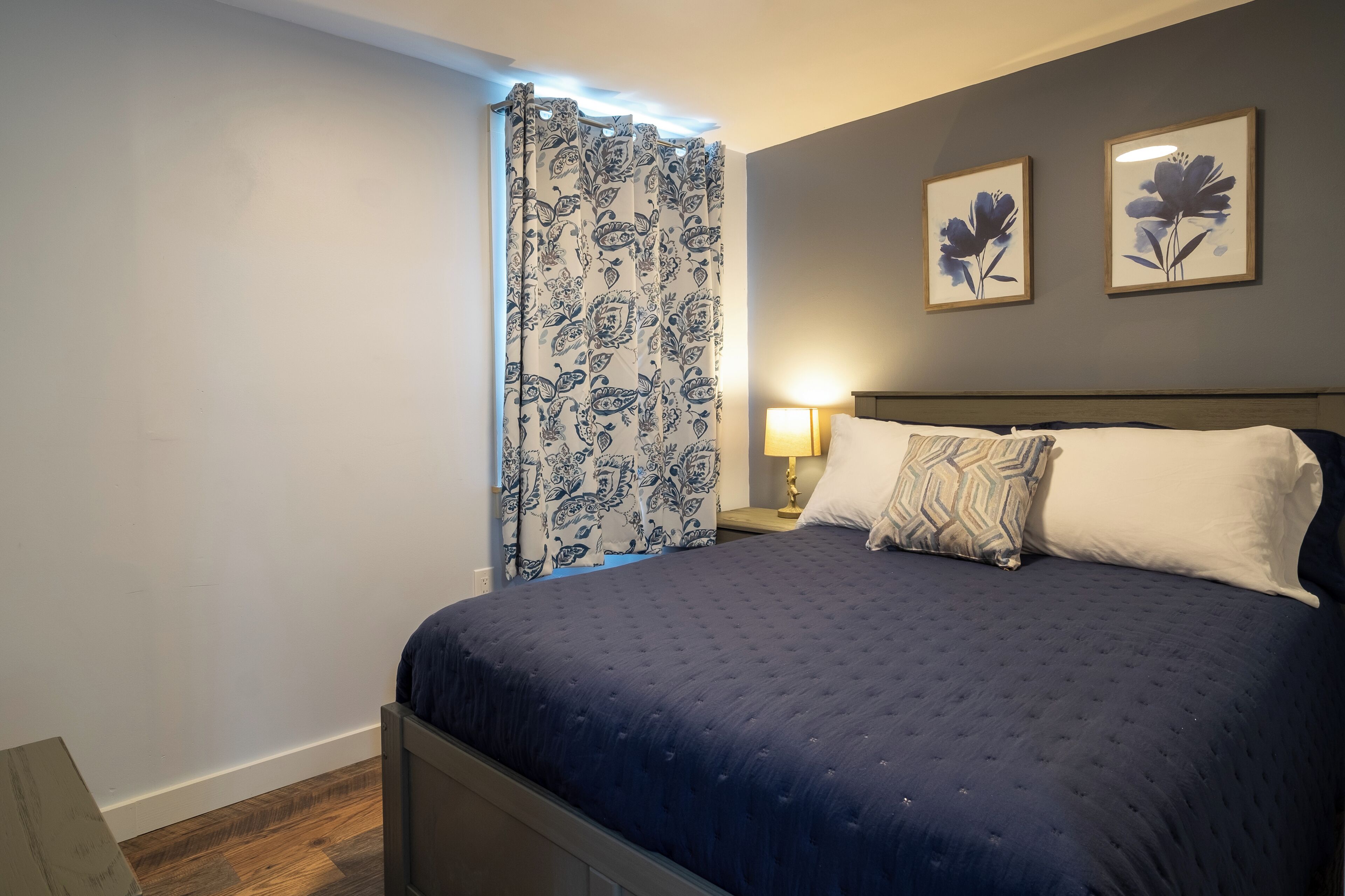 A bedroom with a bed, a nightstand, a lamp, and two framed floral paintings on the wall.