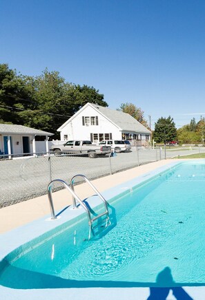 A swimming pool with a metal ladder, a white building with a gabled roof, and a fenced area with parked vehicles. - 3BedroomApt Pool CampFire PlayArea (Bar Harbor)
