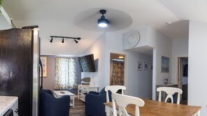 A compact living space with a dining table, two blue chairs, a white chair, a small white table, a television, and a ceiling fan. - 3BedroomApt Pool CampFire PlayArea (Bar Harbor)