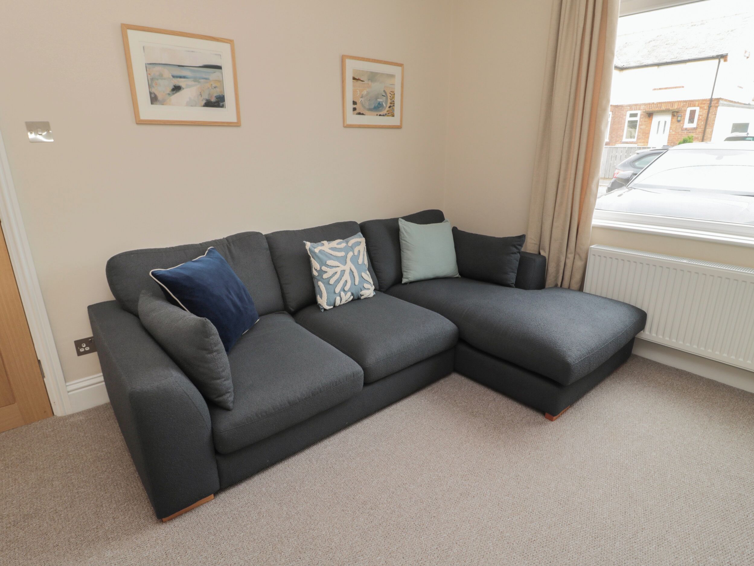 A grey sectional sofa with a blue and white patterned pillow, a light green pillow, and a dark grey pillow in a room with beige walls and a window.