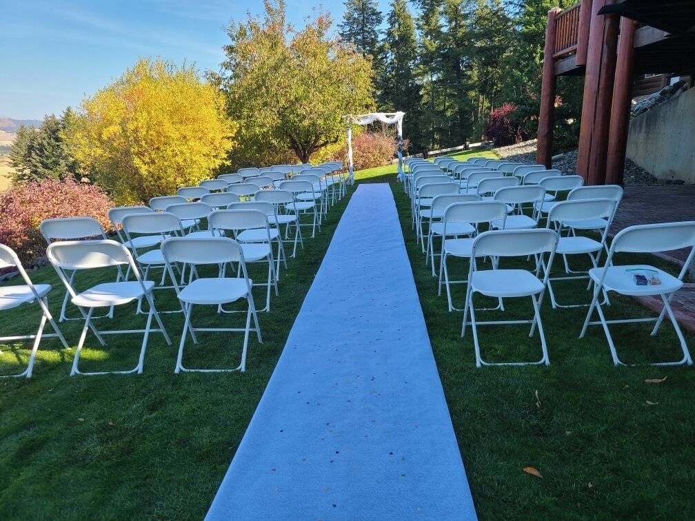 A row of white folding chairs arranged in a semi-circle on a grassy lawn with a blue carpet leading to a white archway.
