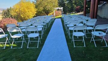 A row of white folding chairs arranged in a semi-circle on a grassy lawn with a blue carpet leading to a white archway.