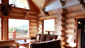 Interior de una cabaña de troncos con un sofá y una mesa de madera, y vistas al exterior.