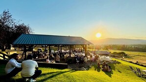 Un área de comedor al aire libre con un pabellón cubierto, mesas y sillas, frente a un atardecer sobre un paisaje montañoso.