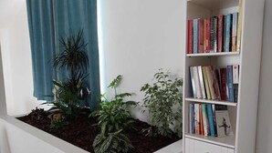 A room with a white bookshelf filled with books, a window with blue curtains, and a planter with various plants. - With a view of Serra de Aire e Candeeiros, with a calm location (Petfriendly). (Cruz da Légua)