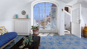 Una habitación con piso azul estampado, un radiador blanco, un sofá azul, una mesa de centro de madera con una planta y un amplio ventanal de arco con barrotes metálicos.