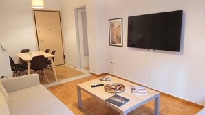 Een moderne woonkamer met een bank, een salontafel met lekkernijen en tijdschriften, een eethoek met stoelen, en een aan de muur bevestigde televisie.
