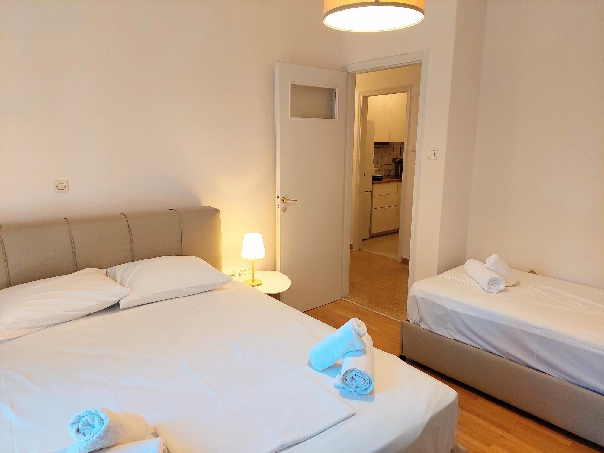 A hotel room with two beds, a bedside table with a lamp, and a view of the kitchen through an open door.
