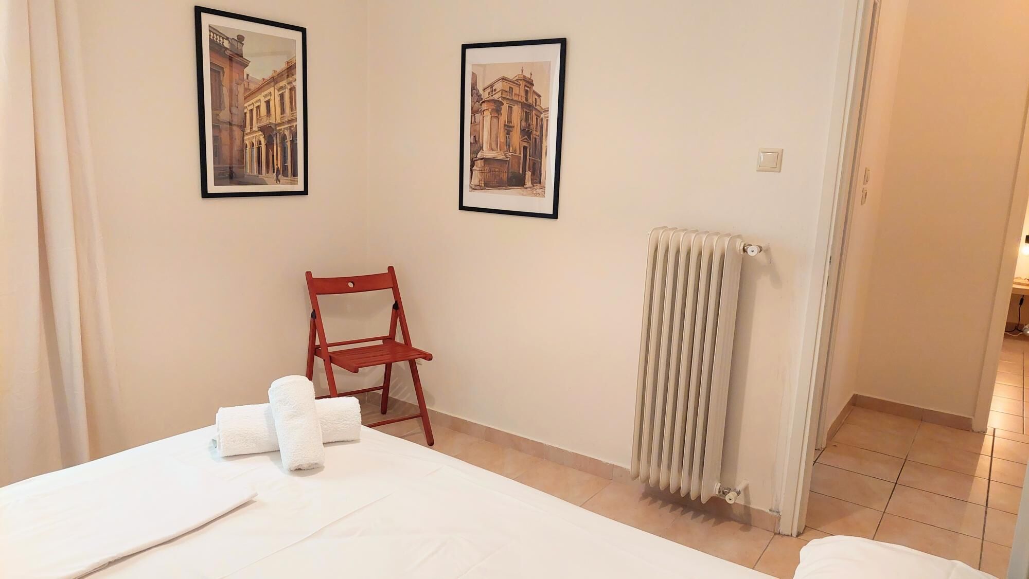 A hotel room with a bed, two framed pictures on the wall, a red chair, and a radiator.