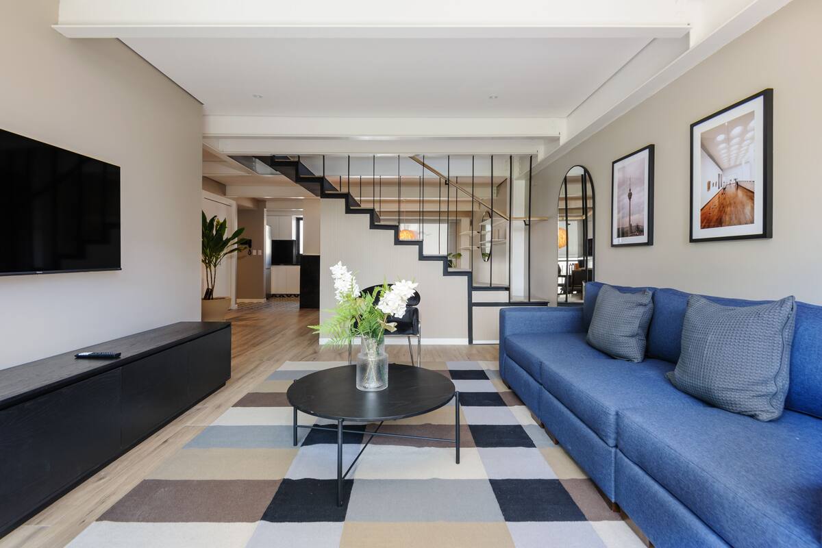 A modern living room with a blue sofa, a black coffee table with a vase, a flat-screen TV, and a staircase leading to another room.