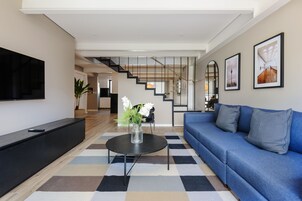 Un salon moderne avec un canapé bleu, une table basse noire ornée d’un vase, un téléviseur à écran plat et un escalier menant à une autre pièce.