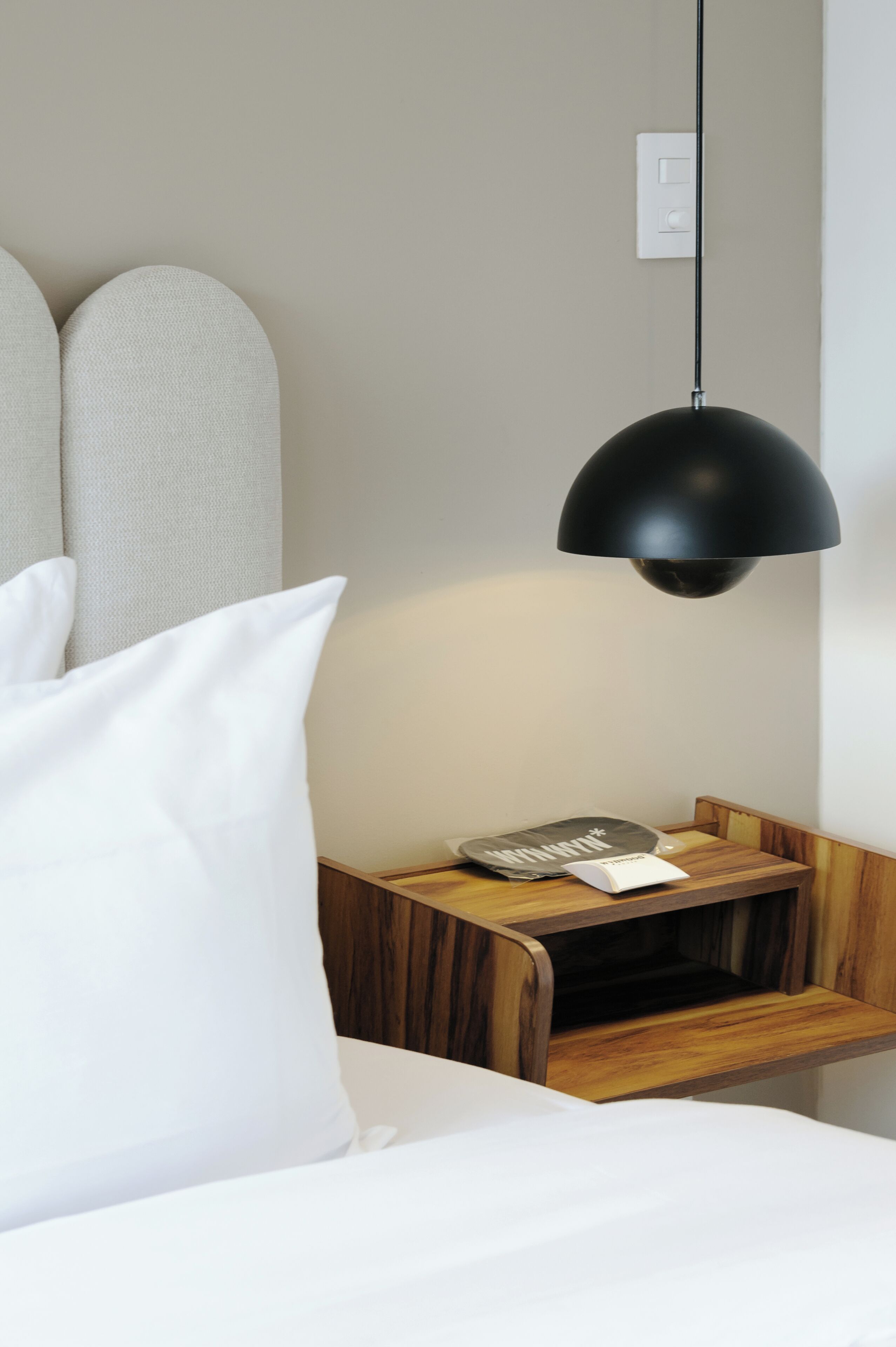 A neatly made bed with white bedding, a black pendant lamp, and a wooden nightstand with a remote control and a small item on it.