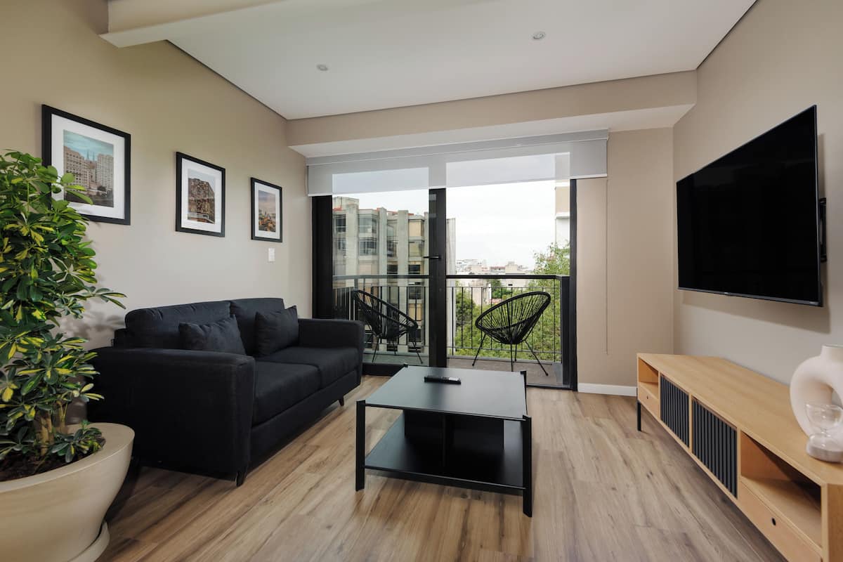 A modern living room with a black sofa, a coffee table, a TV, and a balcony with a view of buildings.