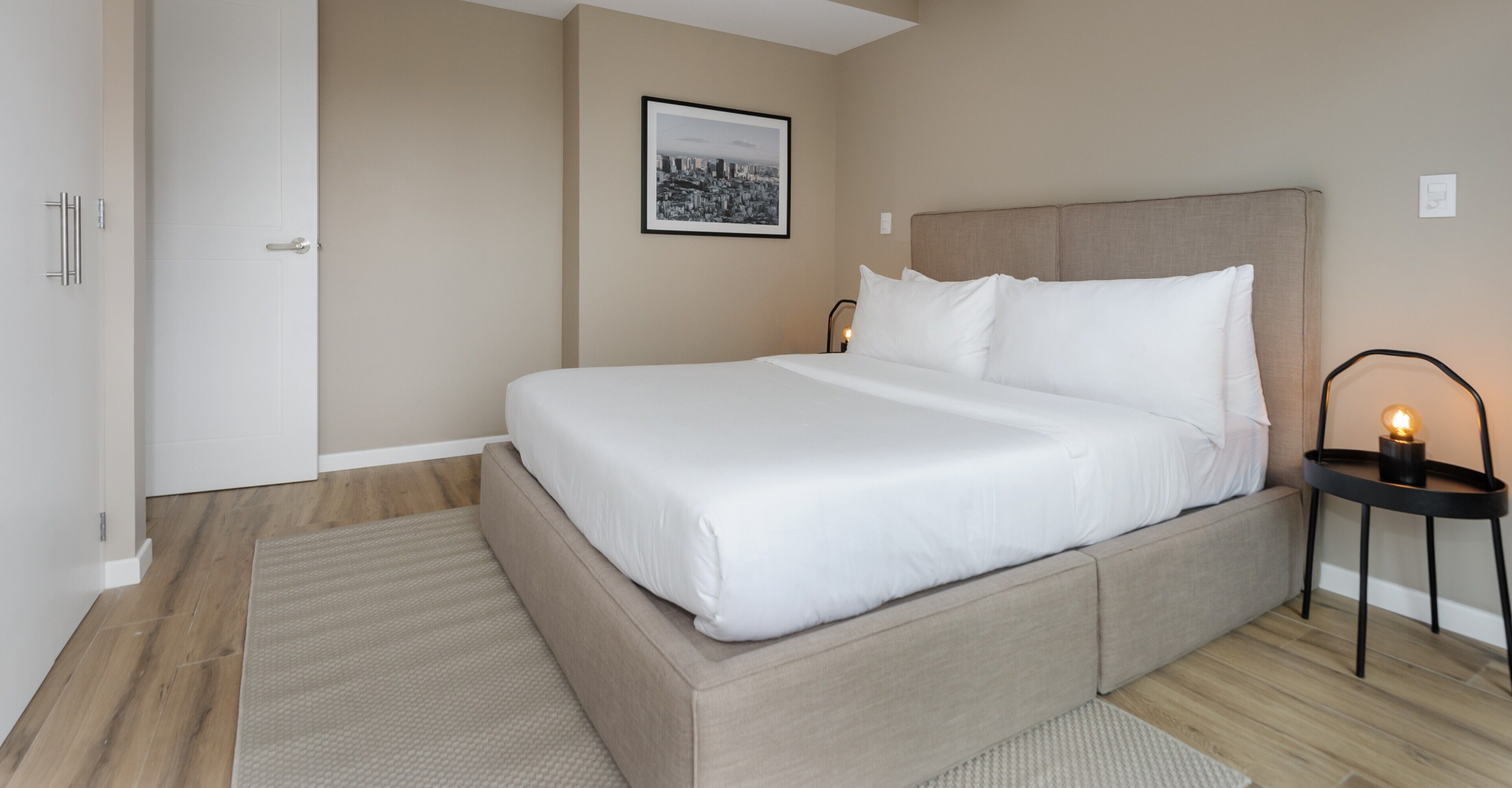 Une chambre moderne avec un grand lit, une tête de lit, une table de chevet avec une lampe et un tableau encadré représentant un paysage urbain accroché au mur.
