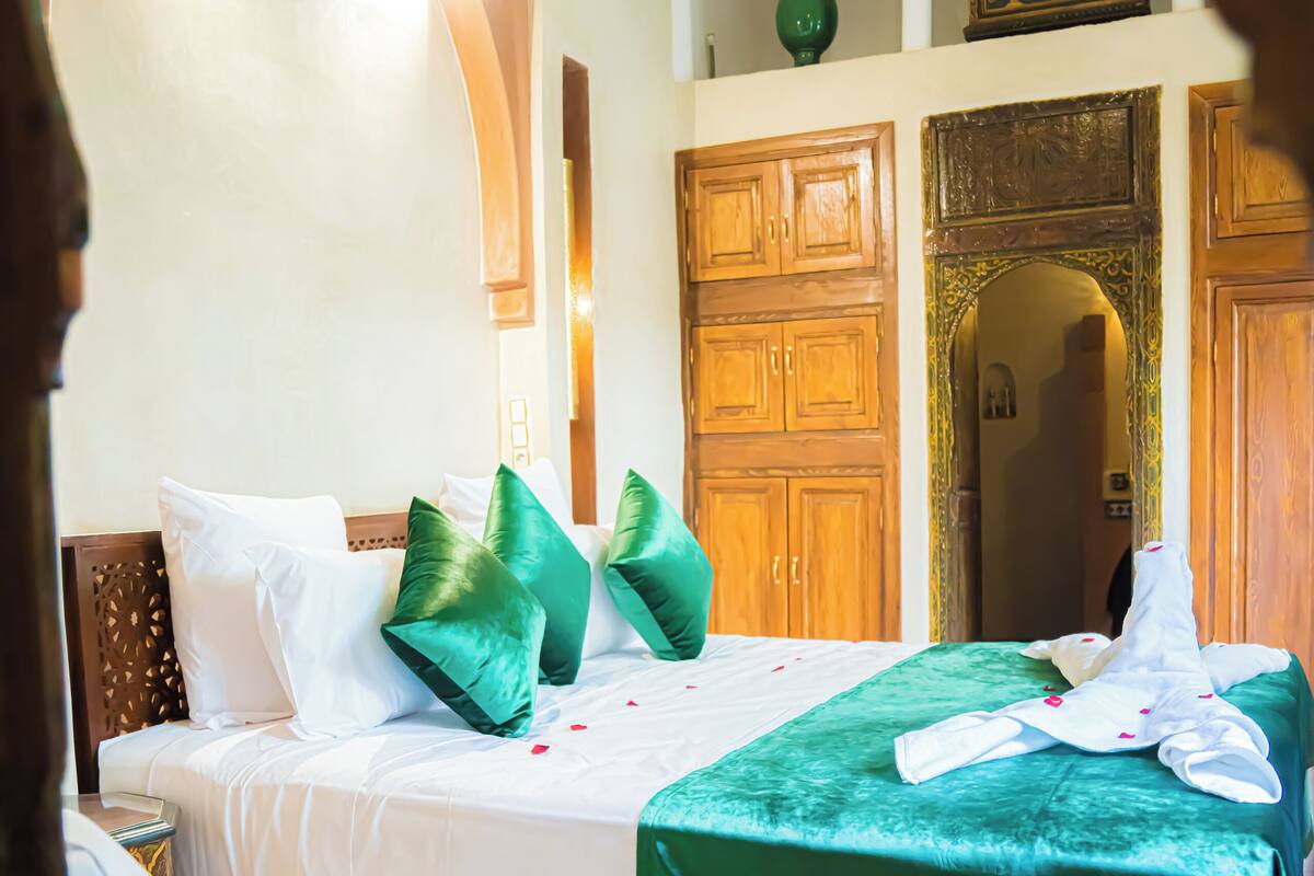 Quarto de hotel com uma cama grande, roupa de cama verde e branca, guarda-roupa de madeira e um tapete estampado.