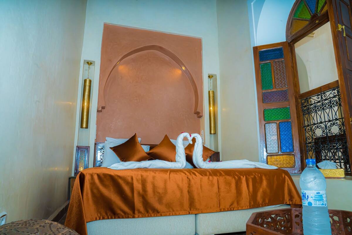 Quarto de hotel com cama, almofadas castanhas, cabeceira decorativa, garrafa de água e janela com vitrais.