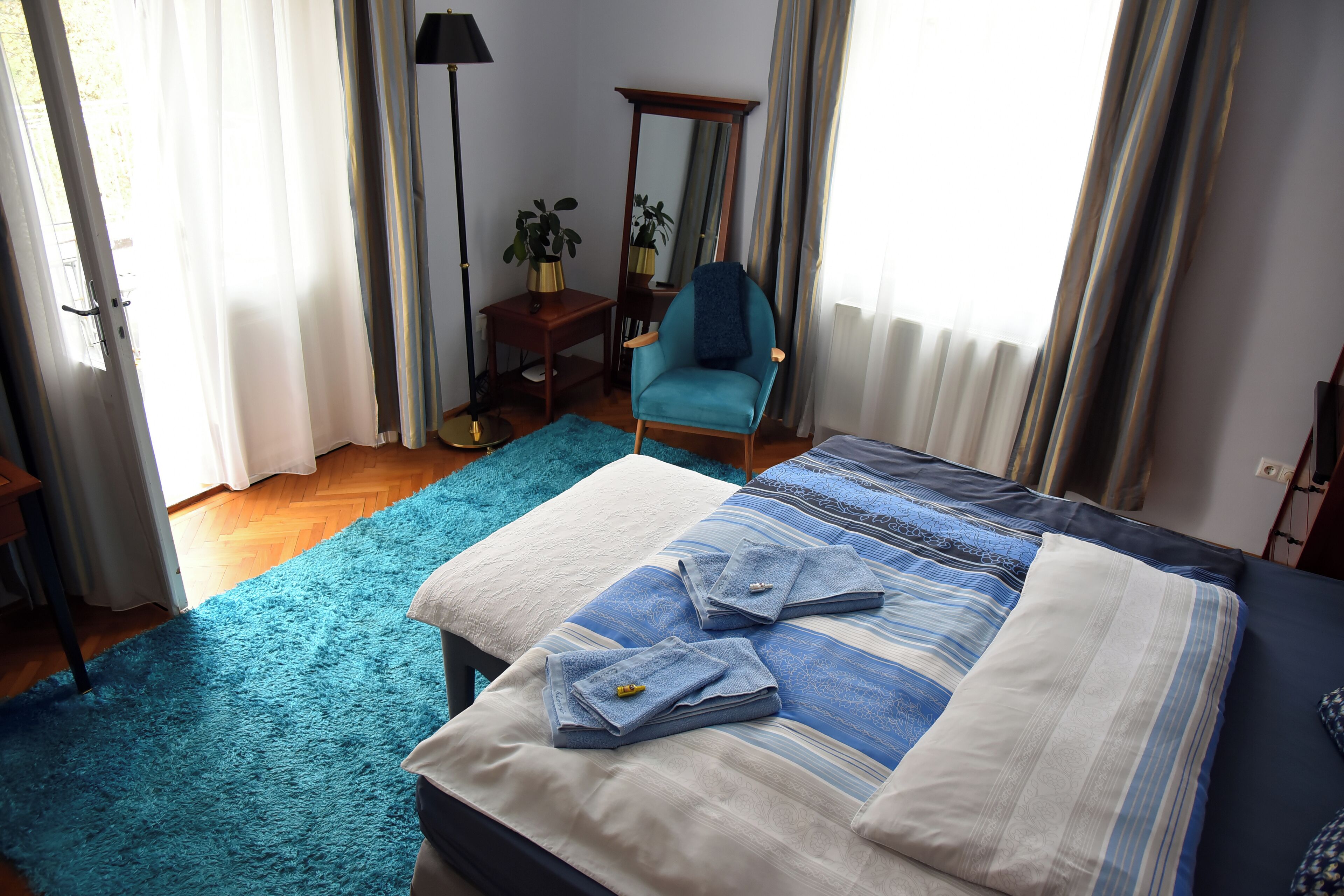 A bedroom with a bed, a chair, a mirror, a small table, and a potted plant.