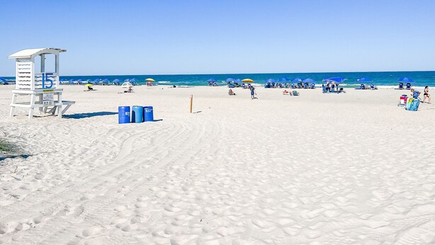 A beach with a lifeguard tower, blue umbrellas, and clear skies. - Cozy Beach Condo Sleeps 6 POOL
 (Carolina Beach)
