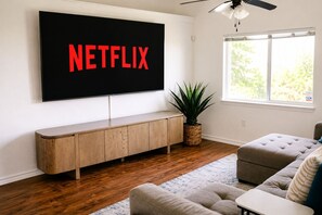 A living room with a flat-screen TV mounted on the wall, a wooden console table, a grey sofa, a potted plant, and a window with a view of greenery. - Family Retreat w/Pool - Golf - Pickleball Sleeps 9 (El Paso)