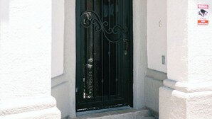 Une porte en métal noir ornée de motifs décoratifs, une porte blanche et un panneau d’avertissement indiquant que les lieux sont sous surveillance vidéo 24 heures sur 24.