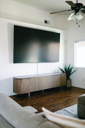 A living room with a large flat-screen TV mounted on the wall, a wooden console table, a potted plant, and a ceiling fan.