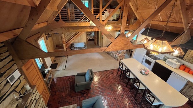 A spacious loft-style room with a high ceiling, exposed wooden beams, a cozy seating area with a sofa and chairs, a kitchen with modern appliances, and a view of the upper floor. - Private, cozy cabin on 13 acres with two ponds (Bloomfield)